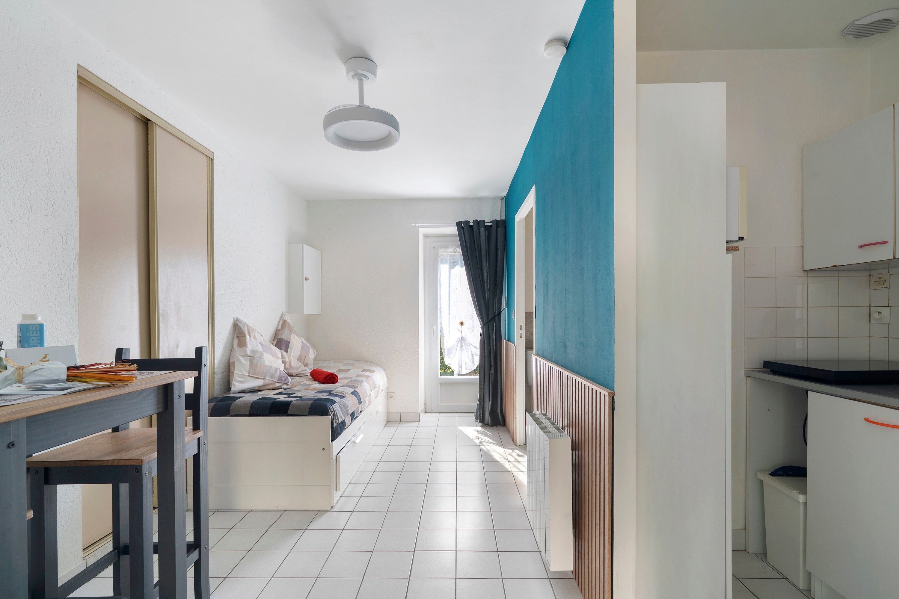 A narrow hallway with a bed, a table, and a kitchenette.