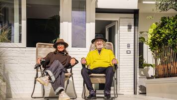Deux personnes sont assises sur des chaises à l’extérieur, devant un bâtiment à la façade blanche et doté d’une porte.