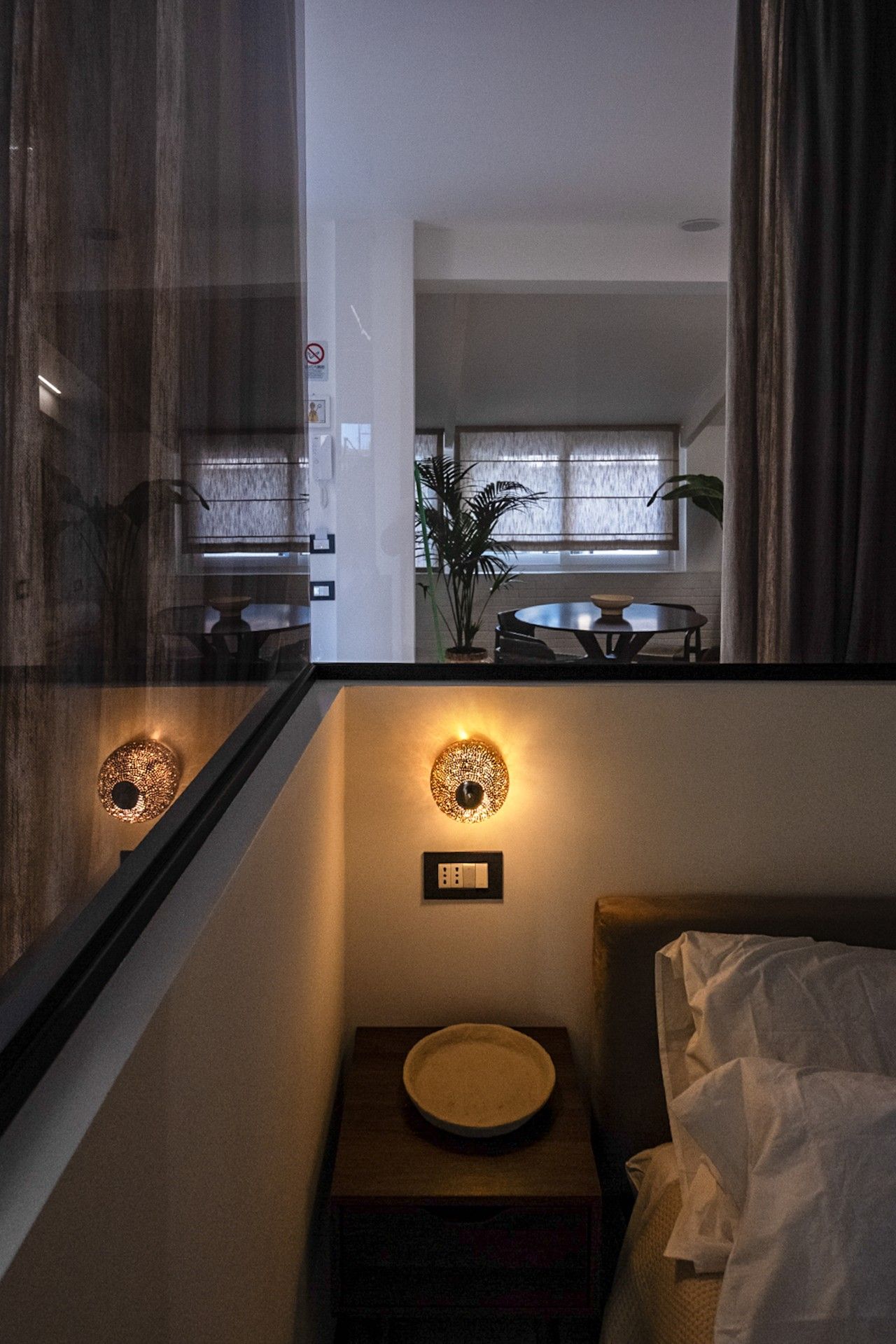 Une chambre d’hôtel avec un lit, une table de chevet sur laquelle se trouve un bol, un luminaire fixé au mur et une fenêtre avec des rideaux.