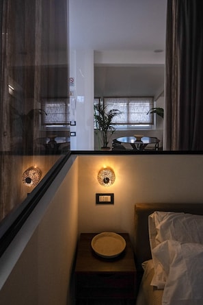 Una camera d'albergo con un letto, un comodino con una ciotola, una lampada a parete e una finestra con tende.