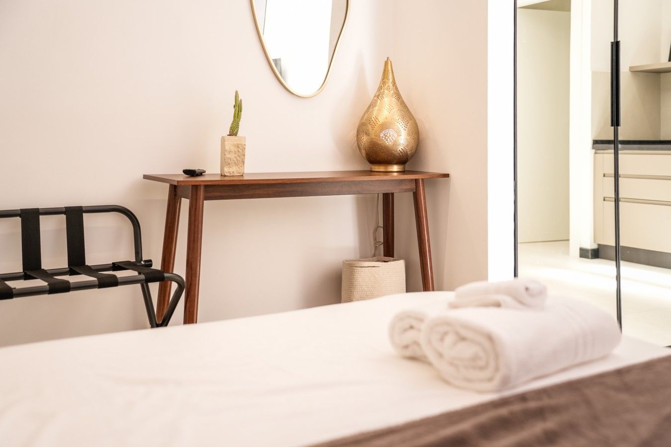 Un letto rifatto con cura, una sedia in metallo nero, un tavolino in legno con un cactus e un vaso decorativo, e uno specchio a parete.