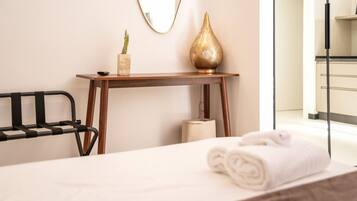 Un lit bien fait, avec une serviette enroulée, une chaise en métal noir, une table d’appoint en bois ornée d’un cactus et d’un vase décoratif, et un miroir accroché au mur.