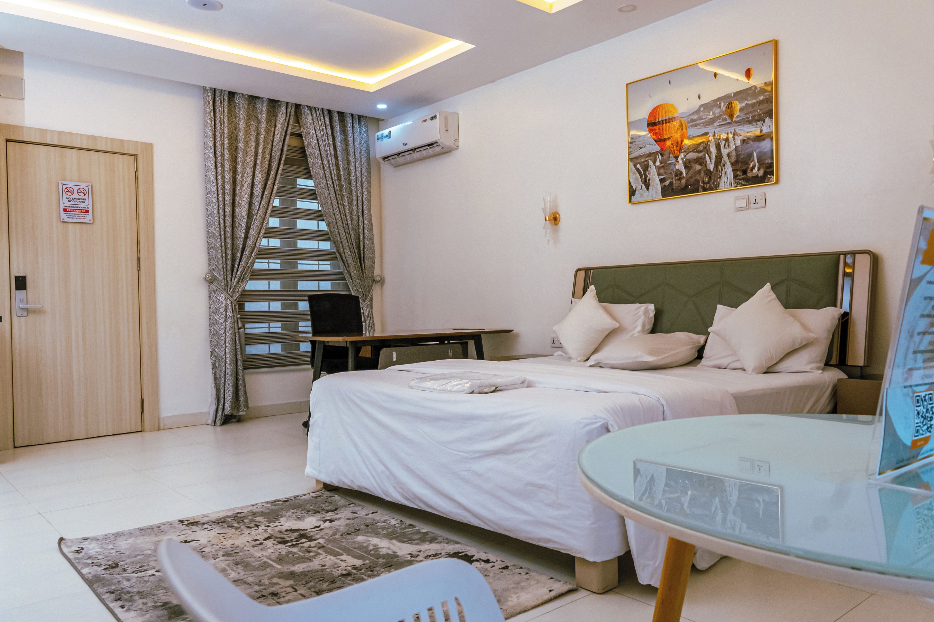 A hotel room with a bed, a desk, a chair, a window with curtains, an air conditioning unit, and a painting on the wall.