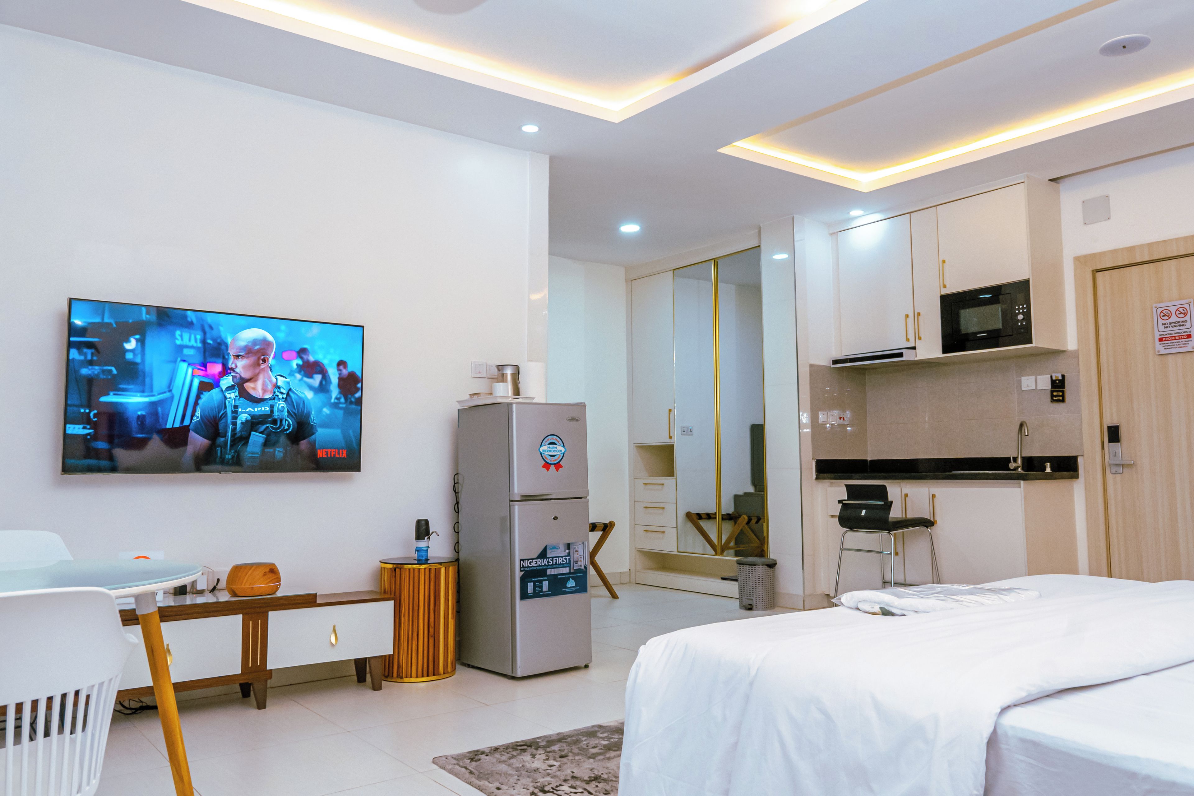 A modern hotel room with a bed, a desk, a television, a minibar, and a kitchen area.
