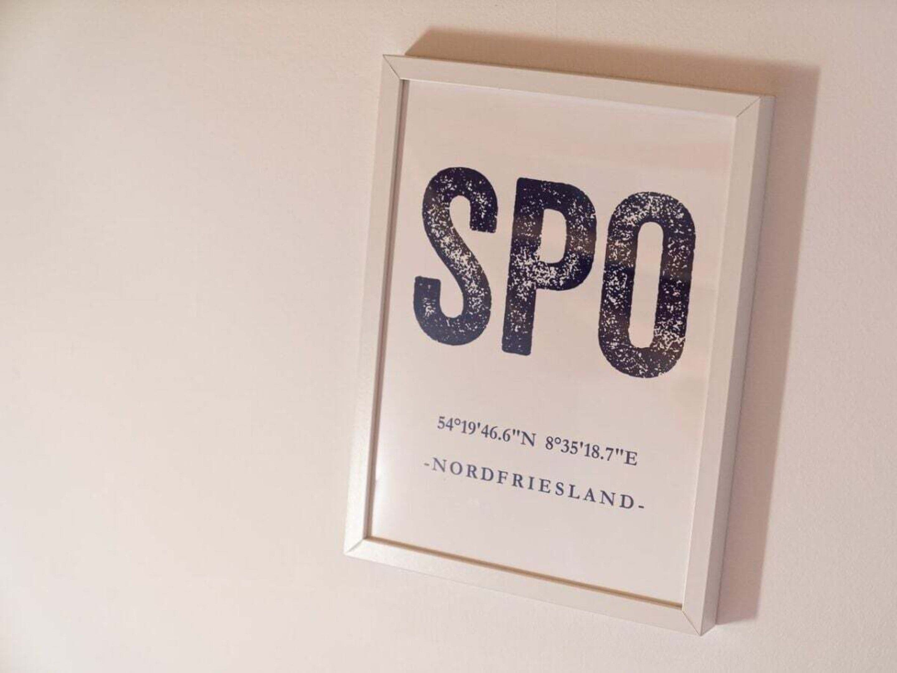 A framed sign with the text 'SPO' and coordinates '54°19'46.6'N 8°35'18.7'E, 'NORDFRIESLAND' below.