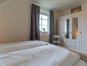 A bedroom with two beds, a window with curtains, a white wardrobe with drawers, and a small chair. - Holiday Home With 1 Bedroom (St. Peter-Ording)
