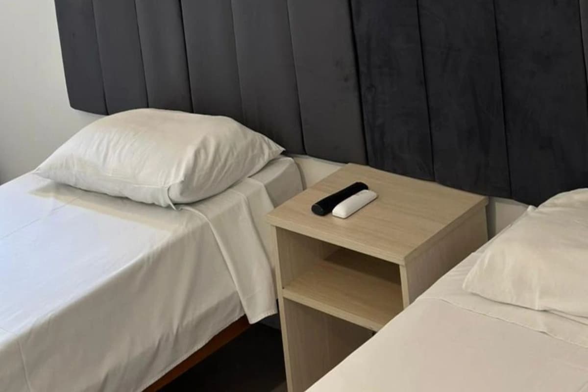 Una cama individual con cabecera, una mesita de noche con control remoto y una almohada blanca.