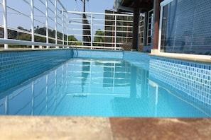 Una piscina azul cristalina con borde de azulejos, rodeada por una cerca blanca y un edificio con fachada de azulejos.