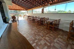 A spacious indoor dining area with wooden tables and chairs, a tiled floor, and large windows offering a view of the outdoors.