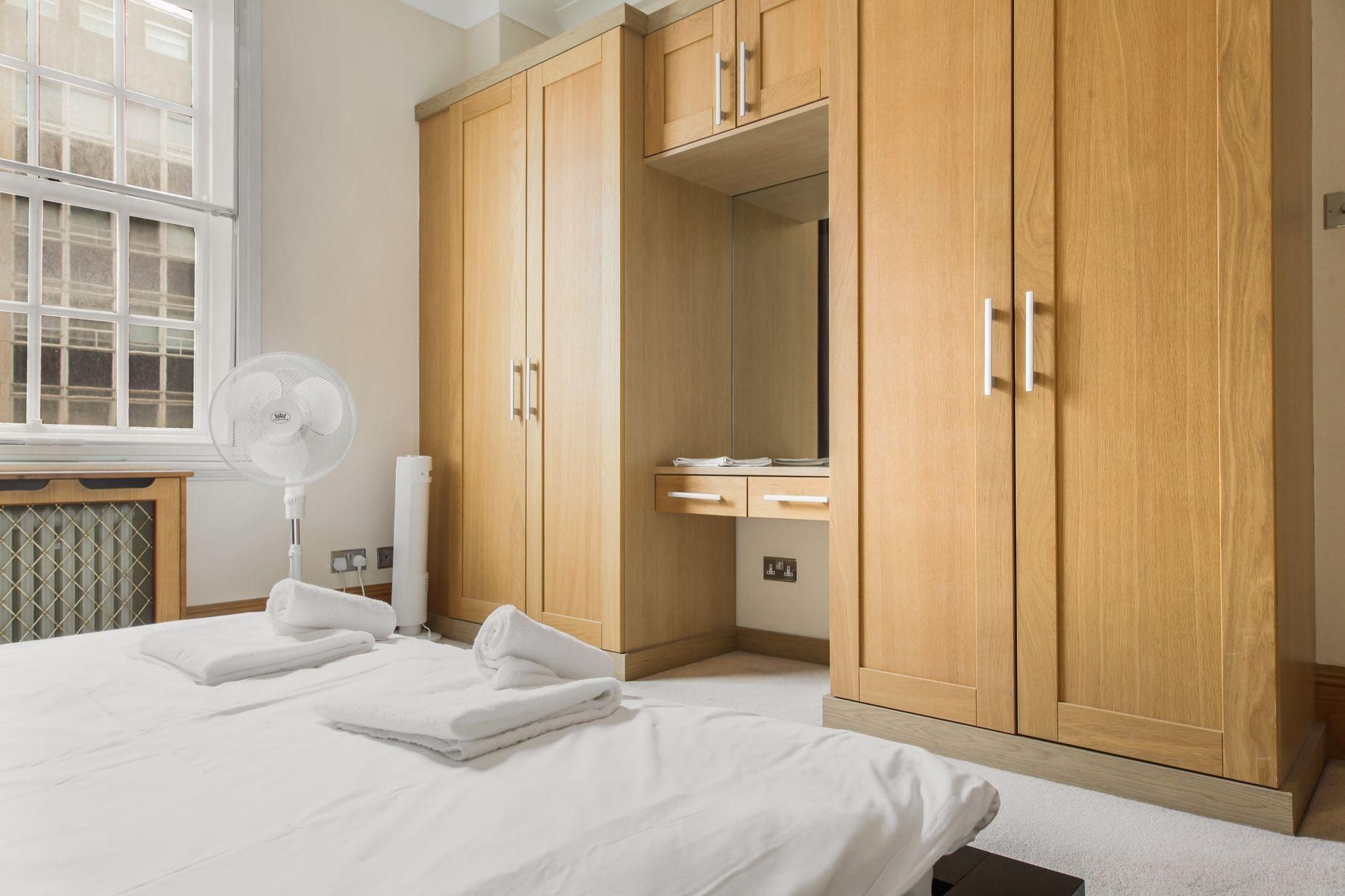 A bedroom with a bed, a fan, wooden wardrobe, and a window.