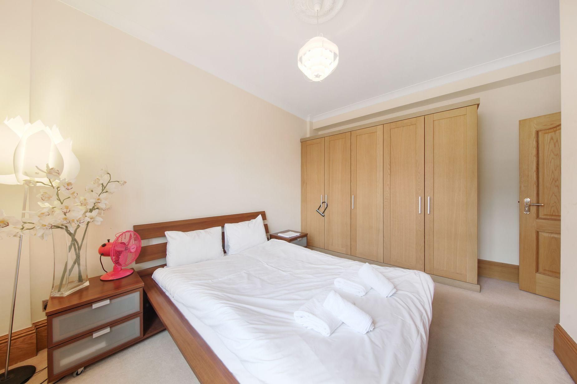 A neatly made bed with white linens, a wooden nightstand with a lamp, a pink fan, and a wooden wardrobe with two doors.
