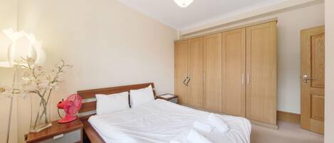 Un lit bien fait, avec du linge de lit blanc, une table de chevet en bois avec une lampe, un ventilateur rose et une armoire en bois à deux portes.