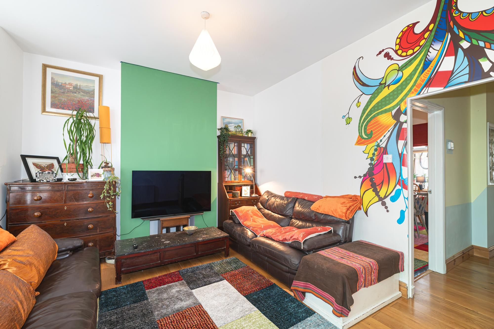 A living room with a television, a sofa, a coffee table, a bookshelf, and a colorful wall mural.
