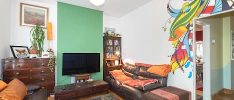A living room with a television, a sofa, a coffee table, a bookshelf, and a colorful wall mural.