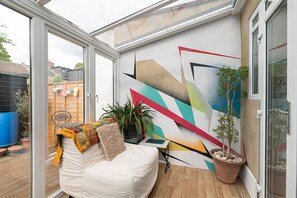 A conservatory with a modern geometric mural, a white sofa with cushions, a small table, and a potted plant. - Stylish & Artistic 3-Bedroom, garden, London Home - Pass the Keys (London)