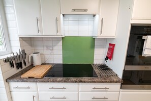 Eine Küche mit weißen Schränken, einer schwarzen Induktionskochplatte, einem Holzschneidebrett und einem roten Feuerlöscher.