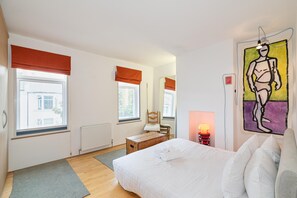 Ein Schlafzimmer mit einem Bett, einer Holzkiste, einem Stuhl, einem Heizkörper und einem Fenster mit Blick auf Gebäude.