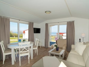 Ein Zimmer mit Esstisch, Stühlen, einer Couch, einem Fernseher und großen Fenstern mit Vorhängen.