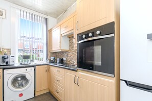 Een kleine keuken met houten kastjes, een inbouwoven, een wasmachine en een raam met zonwering.