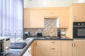 A kitchen with wooden cabinets, a stone backsplash, a black oven, and a dish rack. - Roundhay Retreat – Family-Friendly Home + Parking - Pass the Keys (Leeds)