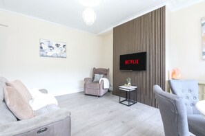 A modern living room with a flat-screen TV mounted on a wall, a grey sofa, a small side table, and a chair. - Roundhay Retreat – Family-Friendly Home + Parking - Pass the Keys (Leeds)