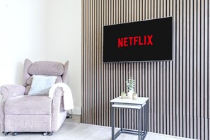 Una sala de estar moderna con un sofá, una mesita con plantas y un televisor de pared que muestra 'NETFLIX'.