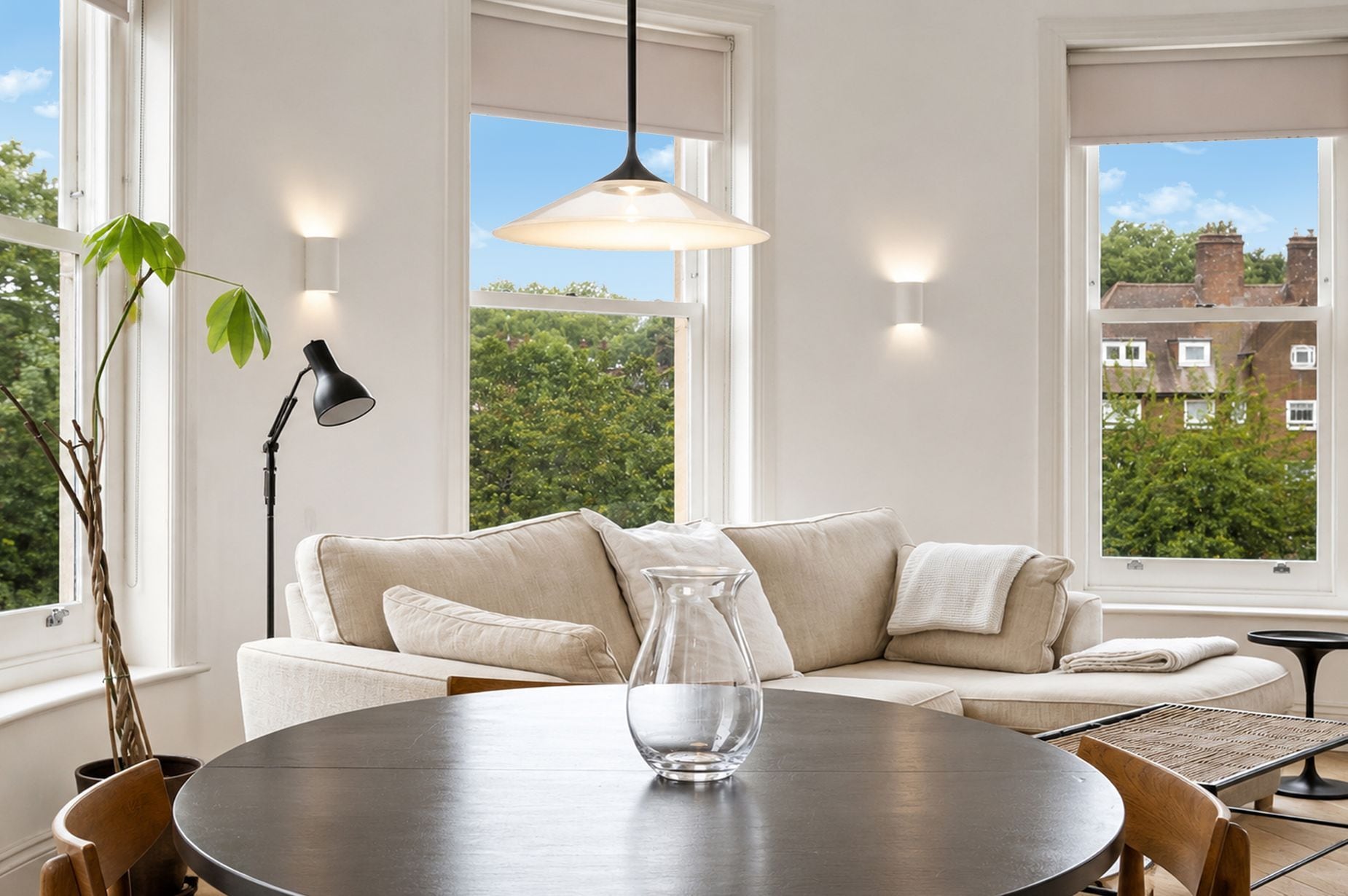 A modern living room with a round dining table, a beige sofa, and a large window offering a view of greenery and buildings.