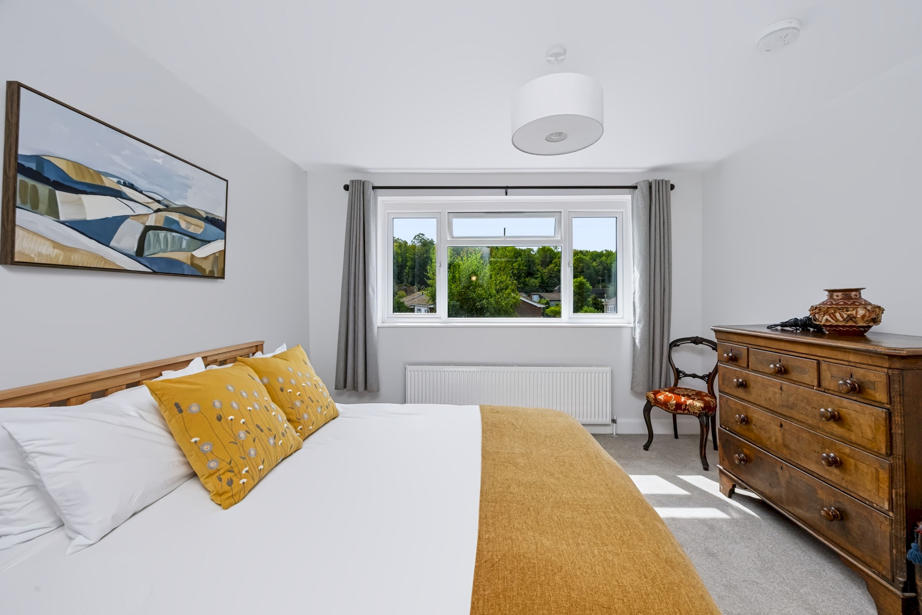 Quarto com cama, cômoda, cadeira, radiador e janela com vista para o verde.
