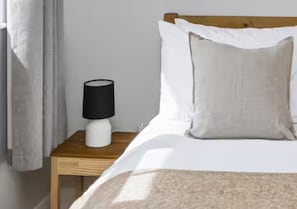 Ein Bett mit weißer Bettwäsche, ein Nachttisch aus Holz mit einer weißen Lampe und ein grauer Vorhang.