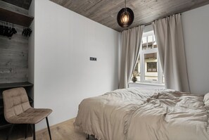 A bedroom with a bed, a chair, a window with curtains, and a hanging light fixture. - Luxury Apt. Downtown Reykjavik - Best Location (Reykjavík)