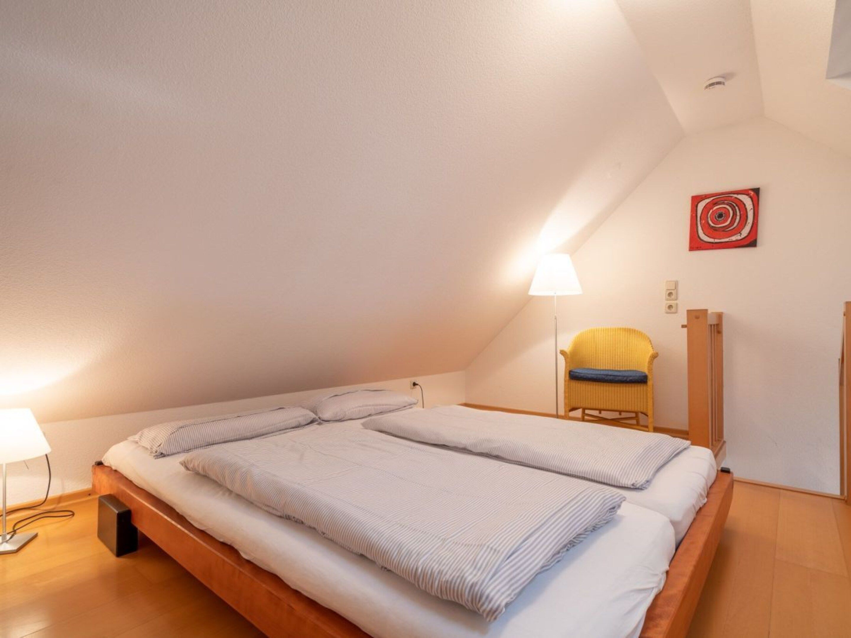 Una cama individual en una habitación con techo inclinado, una lámpara de pie, una silla y una obra de arte colgada en la pared.