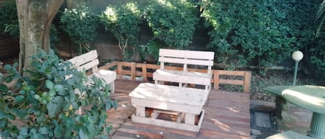 An outdoor seating area with a wooden bench and table, surrounded by greenery.