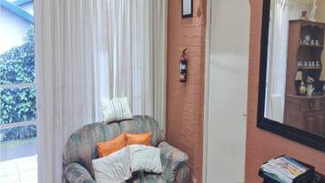 Une pièce avec une fenêtre, un extincteur fixé au mur, une chaise avec des coussins et une table sur laquelle est posée une pile de magazines.