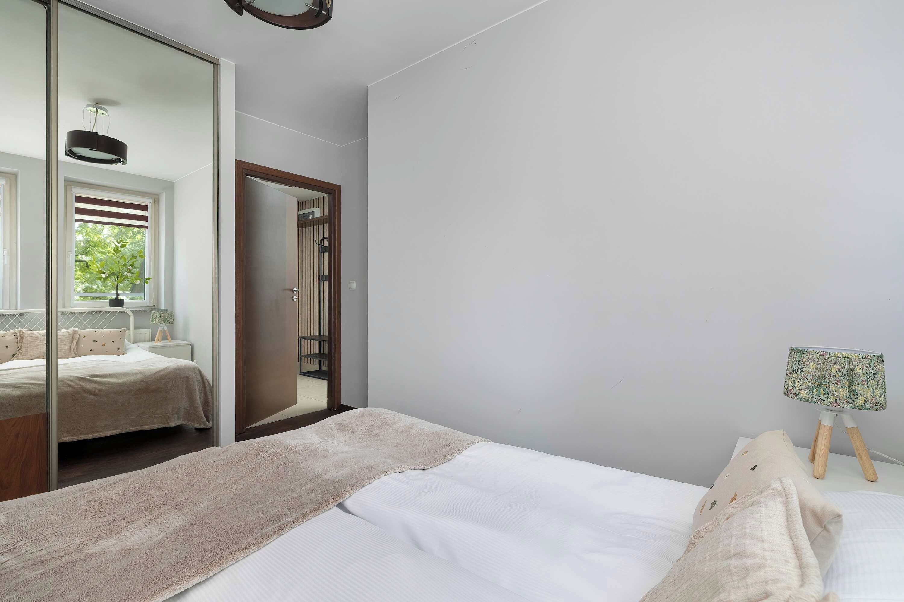 A bedroom with a large mirror, a bed with a white comforter, a bedside table with a lamp, and a small table with a patterned tablecloth.
