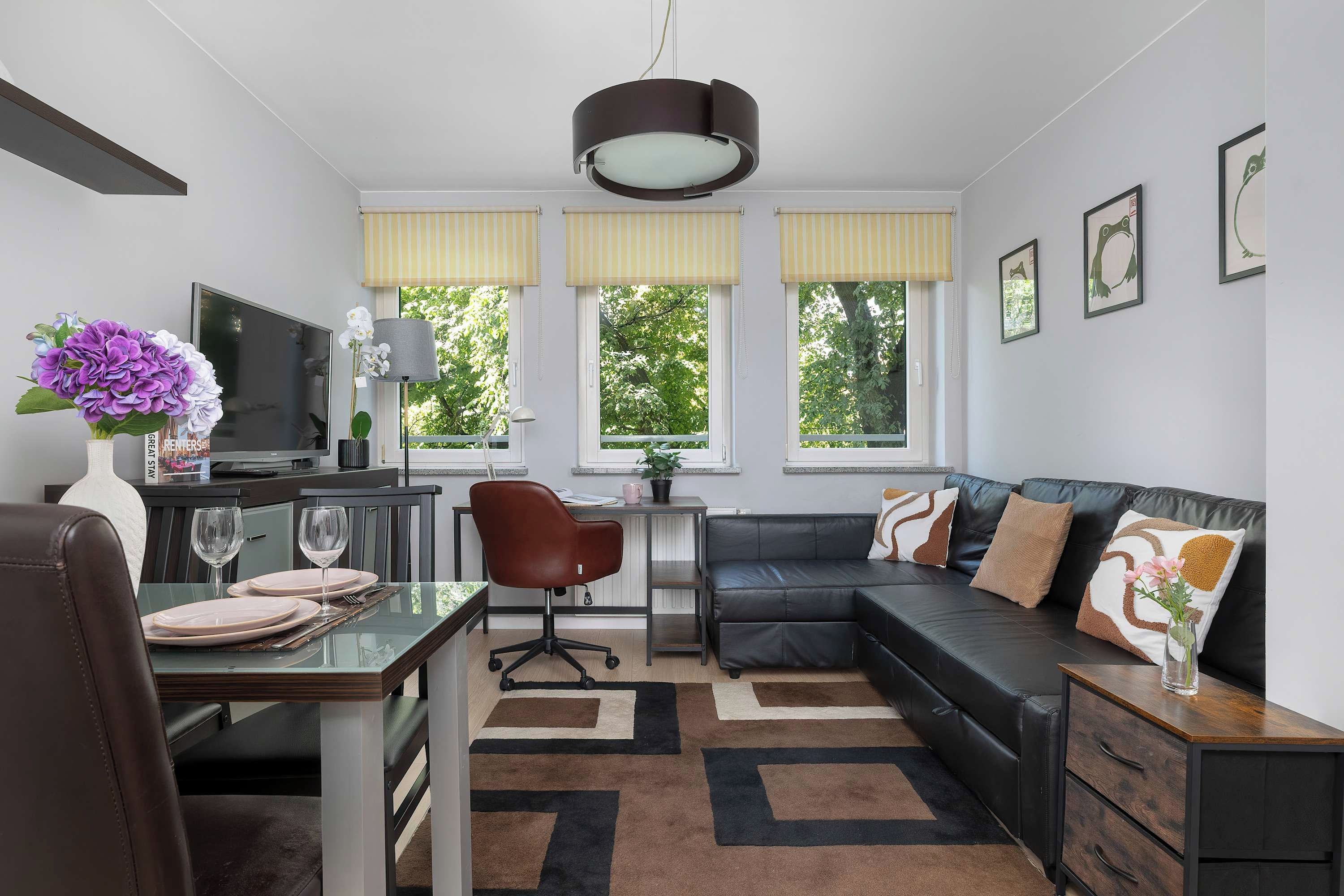 A modern living room with a dining area, a black leather sofa, a TV, and a large window with yellow blinds.