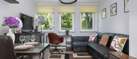 A modern living room with a dining area, a black leather sofa, a TV, and a large window with yellow blinds.