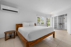 A bedroom with a wooden bed, a small wooden stool, a white air conditioning unit, and a walk-in closet. - Opal Luxury Estate | 6BR Villa, Private Pool & BBQ (Noord)
