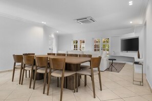 A modern dining area with a round wooden table and six chairs, a white sofa, a TV mounted on the wall, and a window with a view of greenery. - Opal Luxury Estate | 6BR Villa, Private Pool & BBQ (Noord)