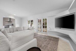 A modern living room with a white sofa, a flat-screen TV mounted on the wall, and a kitchen area visible through an open door. - Opal Luxury Estate | 6BR Villa, Private Pool & BBQ (Noord)