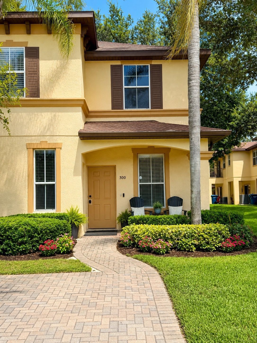 A two-story building with a beige facade, a brown door, and a number '300' on the front. There are two windows with shutters, a small porch with chairs, and a well-maintained lawn.