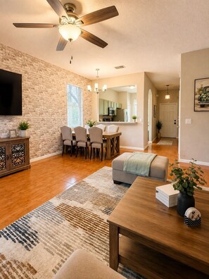 A spacious living room with a wooden dining table, a ceiling fan, and a kitchen in the background. - Spacious Modern Resort Townhome Sleeps 10 with Pools, Lazy River and Near Disney (Davenport)