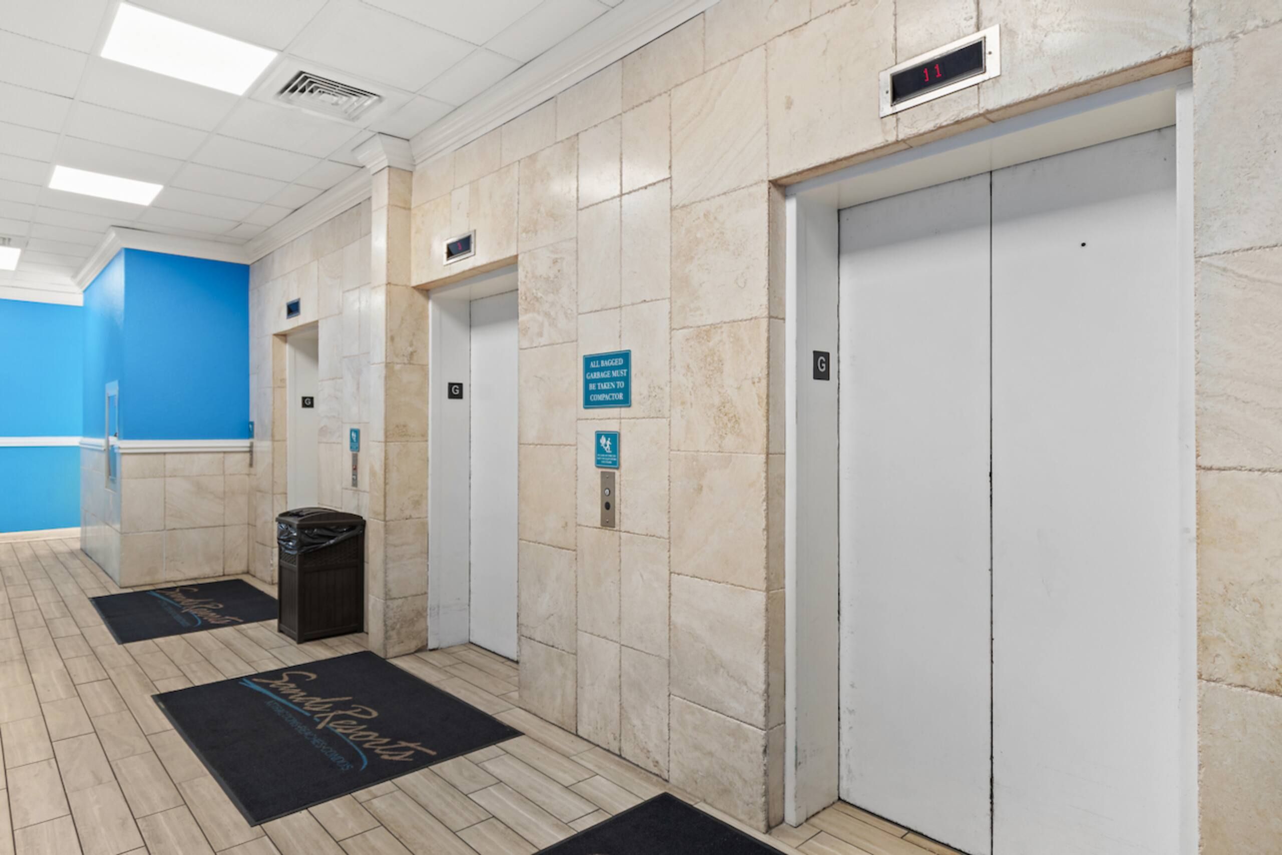 An elevator area with a blue wall, a trash bin, and a doormat with a logo.