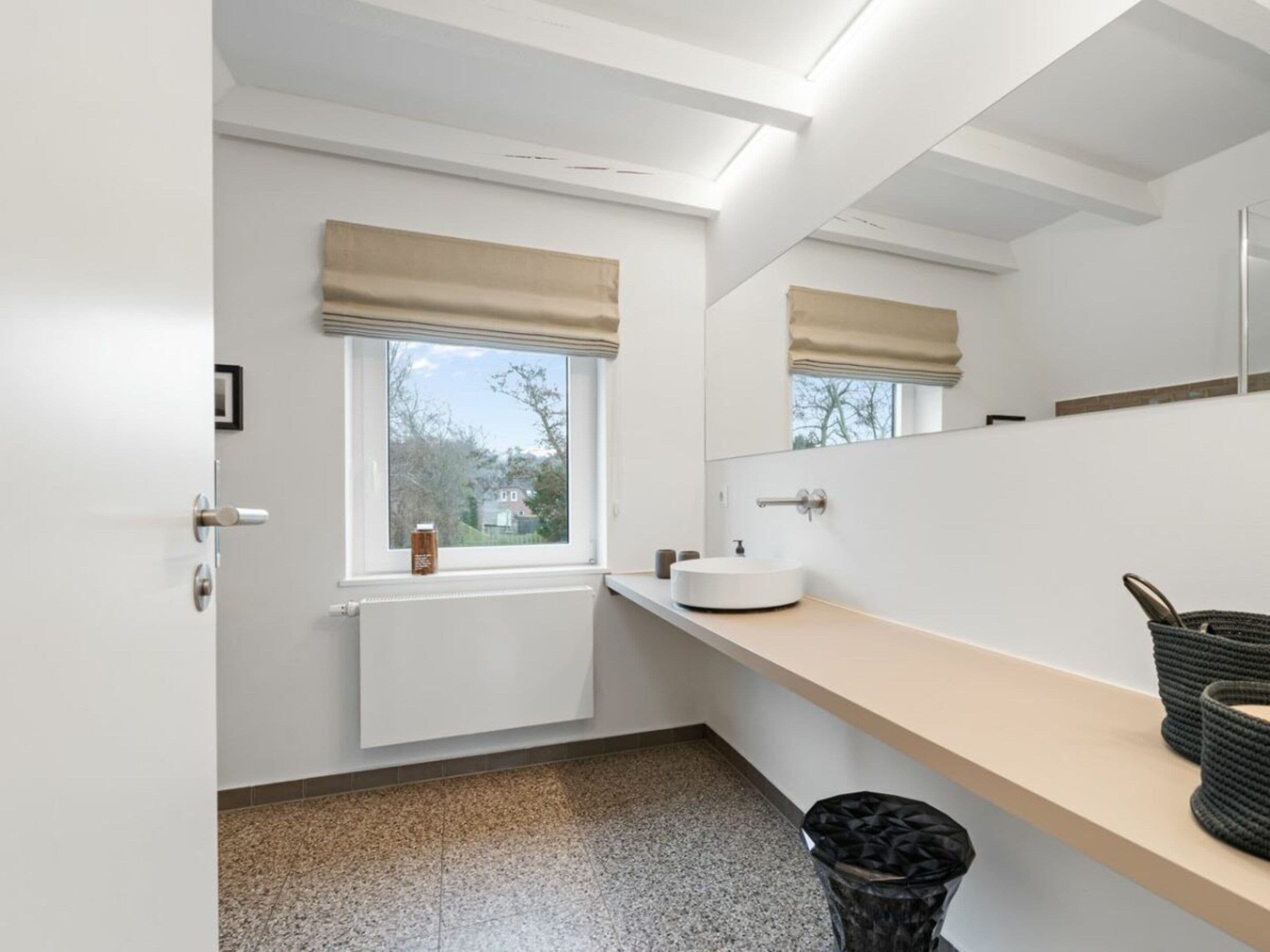 A modern bathroom with a white sink, a mirror, a window with a beige blind, and a black trash bin.