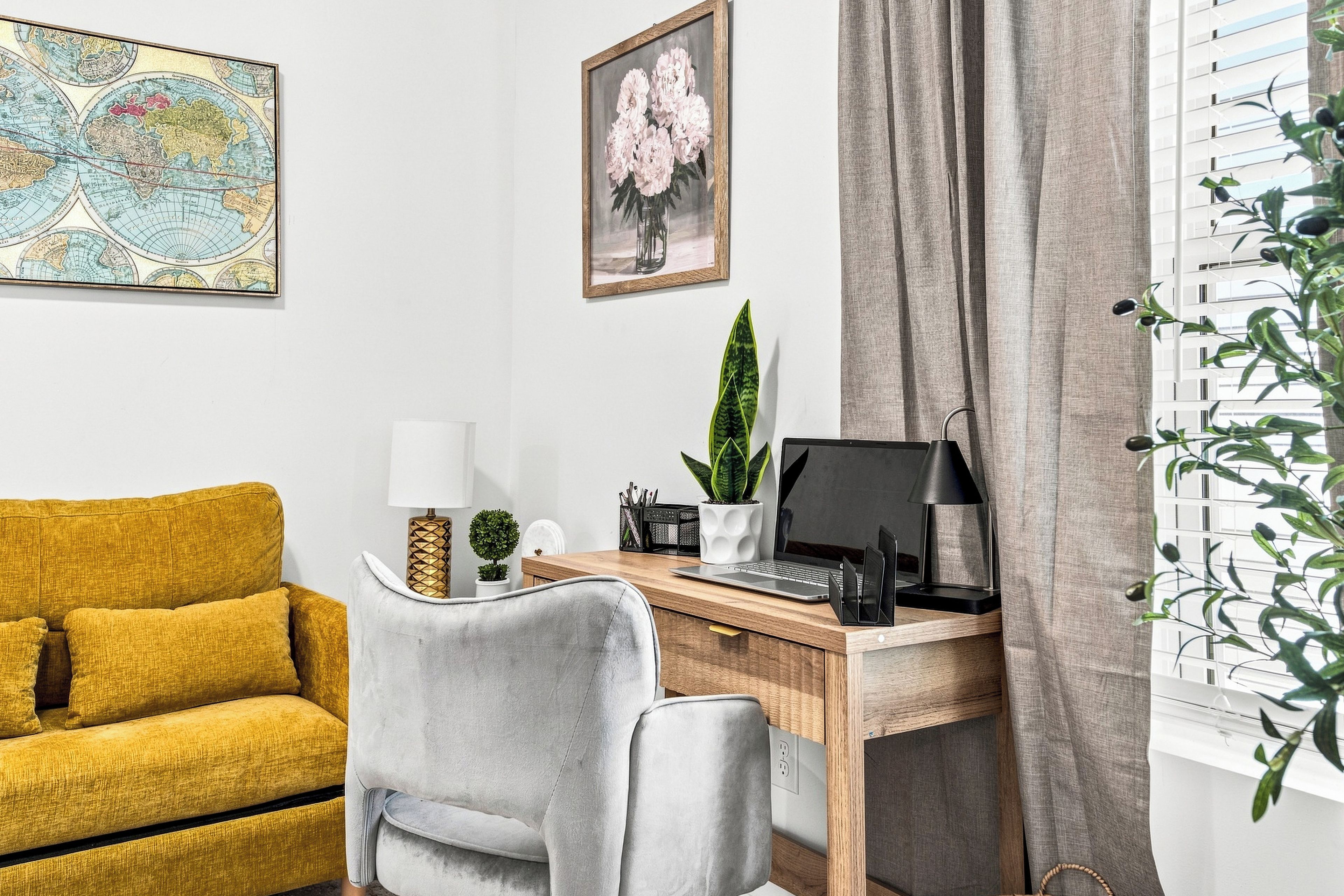 A room with a wooden desk, a yellow sofa, a grey chair, a map, and a flower painting.