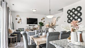 A modern living room with a dining area, featuring a marble countertop, grey upholstered chairs, and a flat-screen TV mounted on the wall. - The Velvet Stay (Granite Quarry)