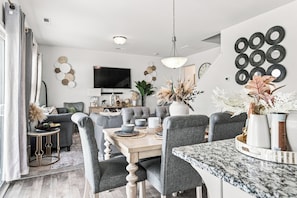 Ein modernes Wohnzimmer mit Essbereich, ausgestattet mit einer Marmorarbeitsplatte, grau gepolsterten Sesseln und einem flachbildfernseher an der Wand.