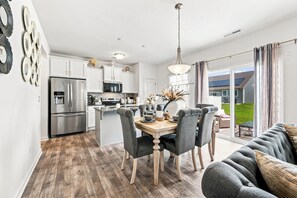Una cocina moderna con electrodomésticos de acero inoxidable, un comedor con sillas tapizadas en gris, y vistas a un jardín verde a través de puertas correderas grandes.