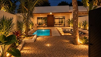 A modern outdoor pool area with a pebble ground, surrounded by tropical plants and a wooden deck.
