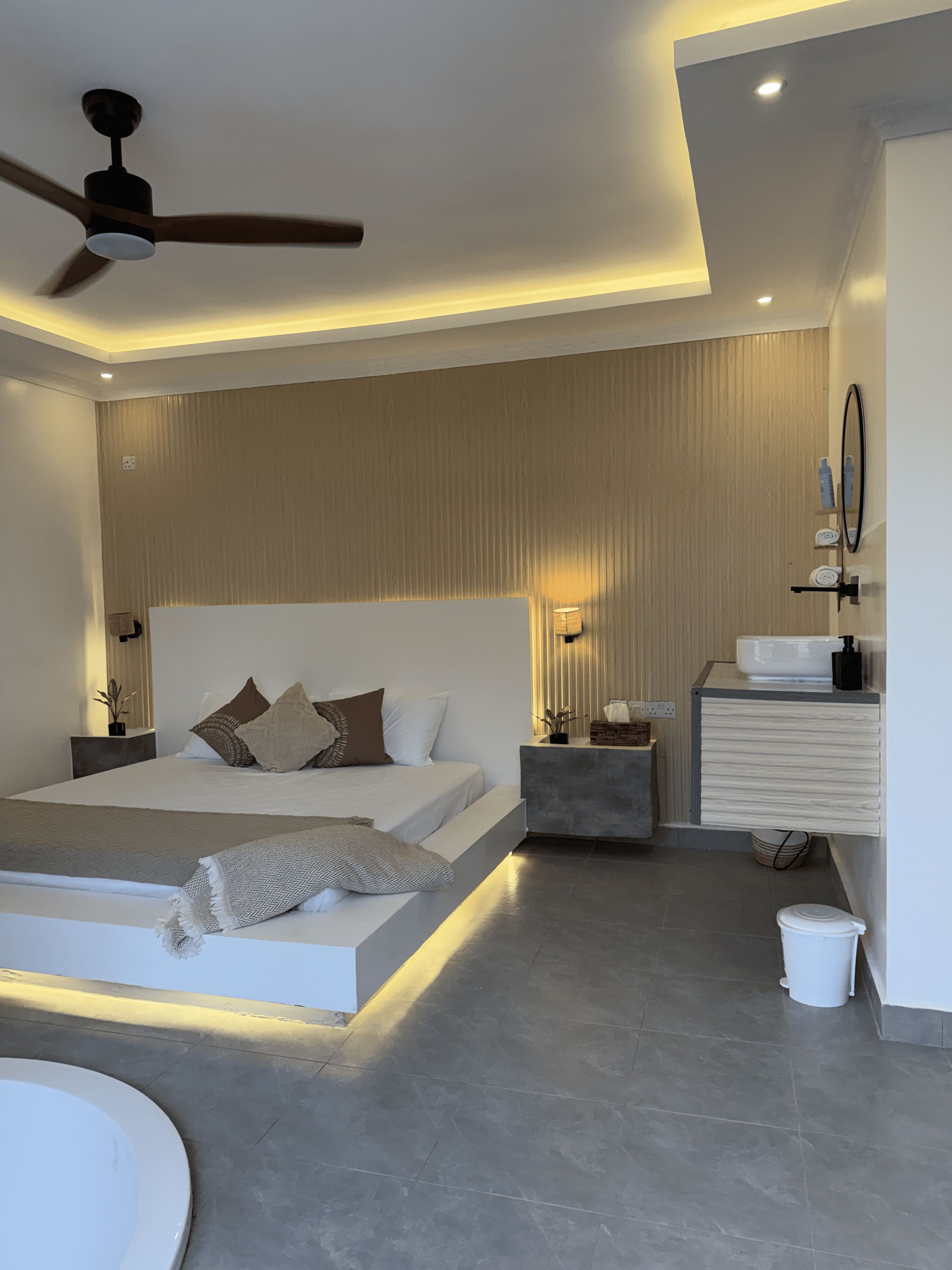 A modern bedroom with a bed, bedside tables, a nightstand, a ceiling fan, and a bathroom with a sink and mirror.