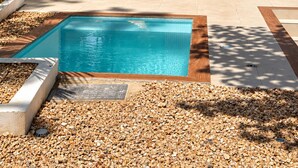 A modern outdoor pool area with a rectangular pool, surrounded by pebbles and a wooden deck. There are two wicker chairs and a table with cushions.